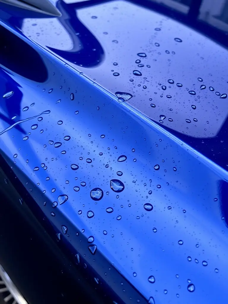 A car with a ceramic coating, water beading on the surface, demonstrating the hydrophobic properties and protective layer. The car should look glossy and well-maintained.